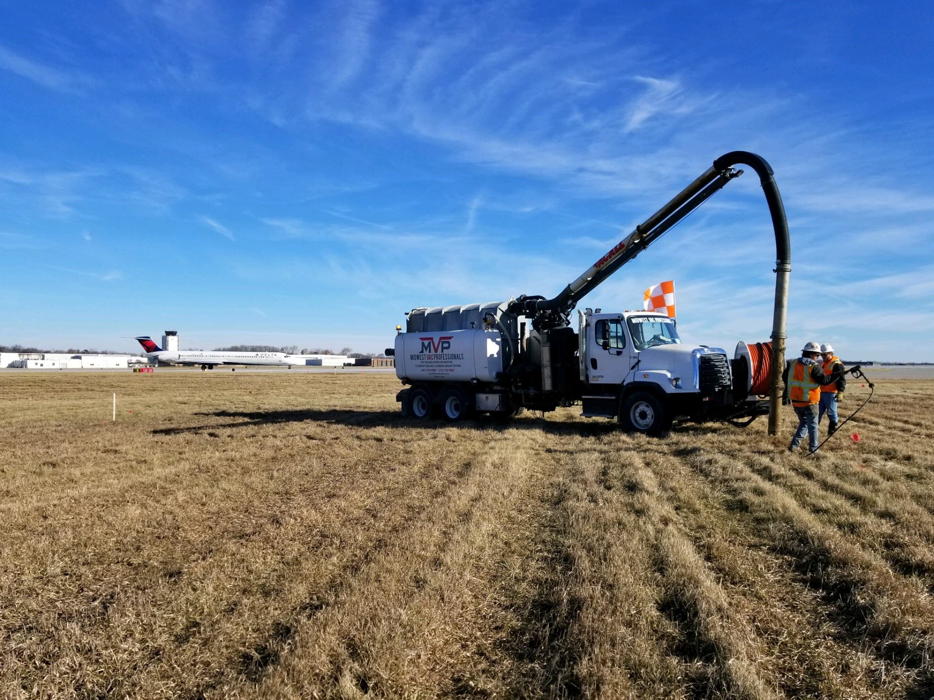 MVP vacuum truck with extended boom hose performing utility work in a field near an airport runway, with two crew members in high-visibility gear and a plane in the background.