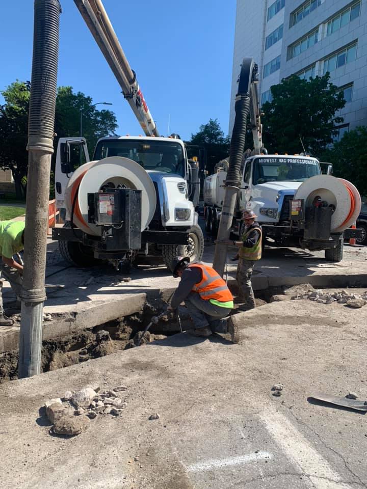 Two MVP vacuum trucks with hose reels working a street excavation site, with crew members in high-visibility gear digging near an exposed utility opening, downtown buildings in the background.